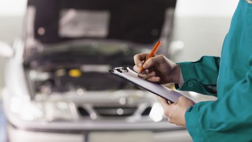 Mechaniker mit Klemmbrett beim Notieren des Arbeitsfortschrittes vor Auto mit geöffneter Motorraumhaube.