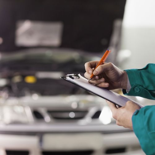 Mechaniker mit Klemmbrett beim Notieren des Arbeitsfortschrittes vor Auto mit geöffneter Motorraumhaube.